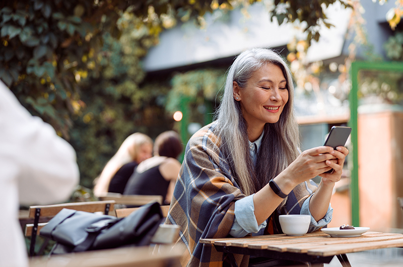 Happy mature Asian woman surfs internet on mobile phone sitting at small table on outdoors cafe terrace on autumn day